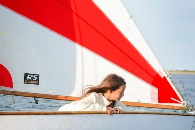 A young sailor leaning over the bow of a dinghy under a red and white sail
