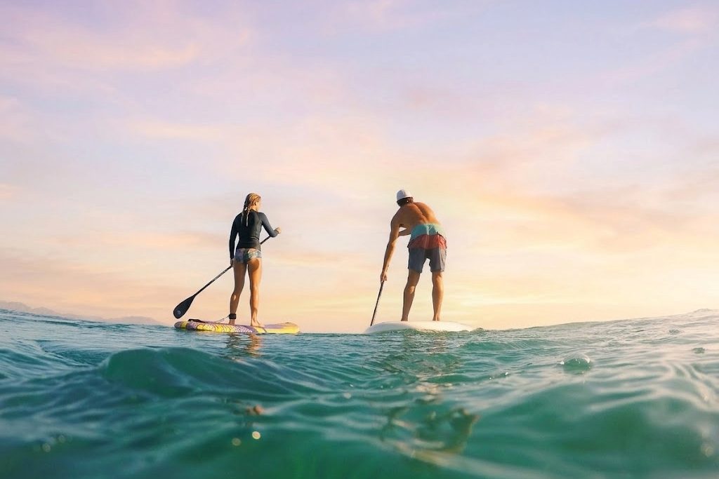 Two paddleboarders crossing calm water under a pink and gold sunset sky